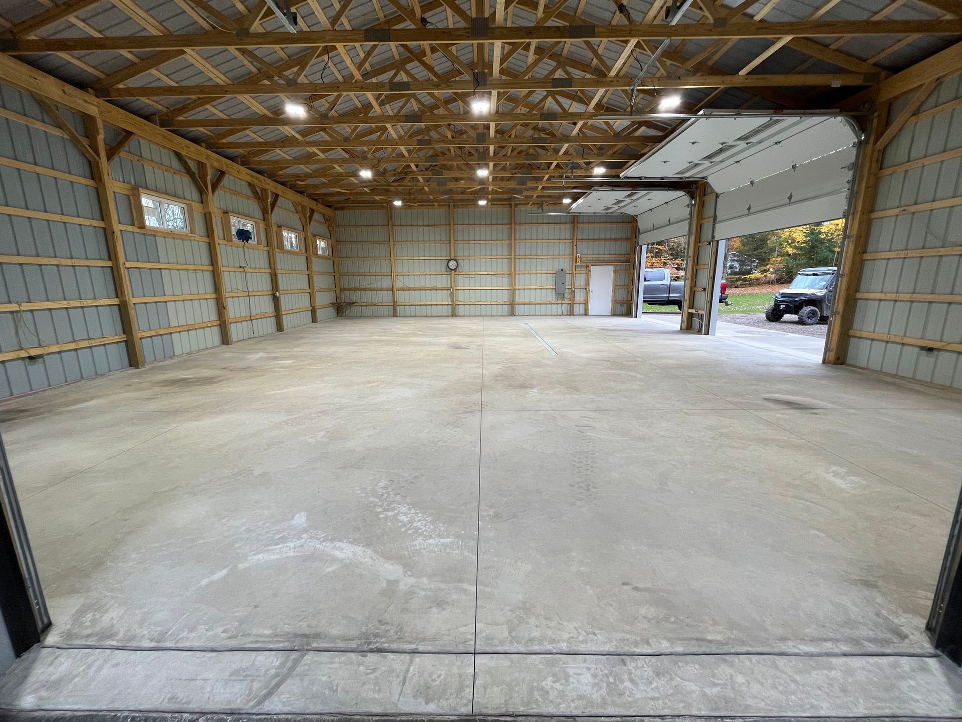Empty concrete floored barn interior with open garage door and overhead lighting.