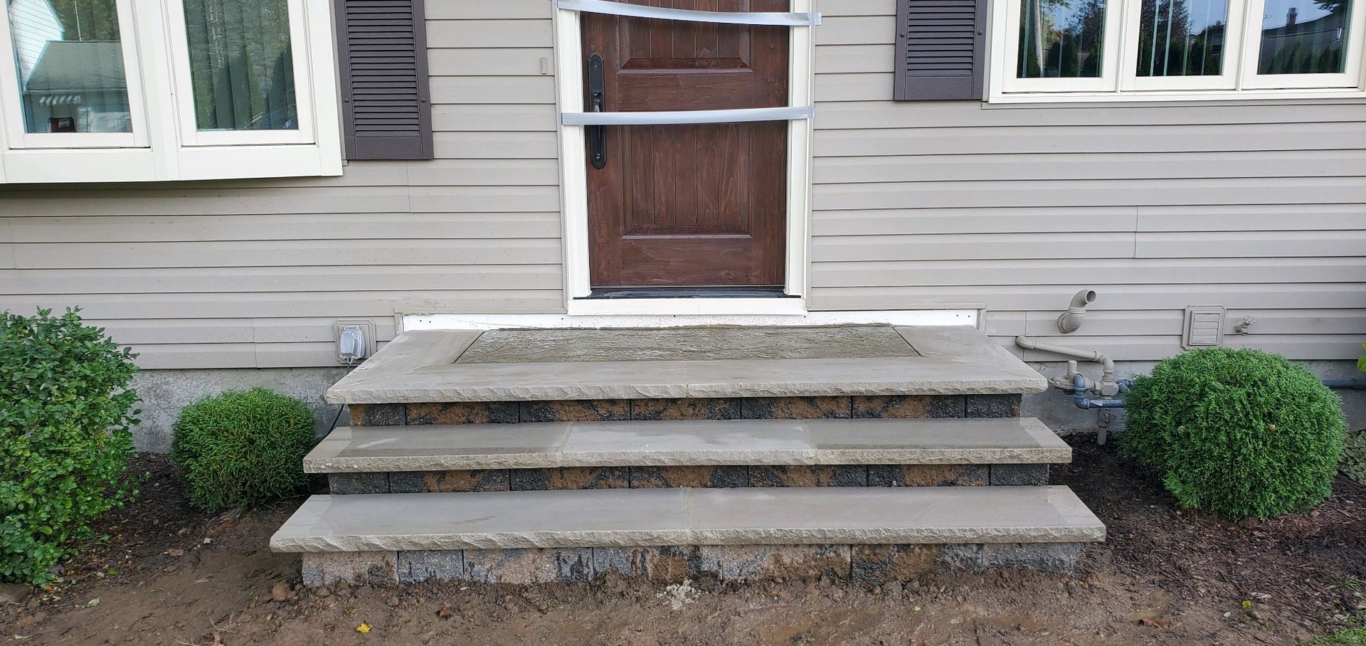 Concrete steps leading to a brown door with a screen door. Green bushes flank the steps on a tan house.
