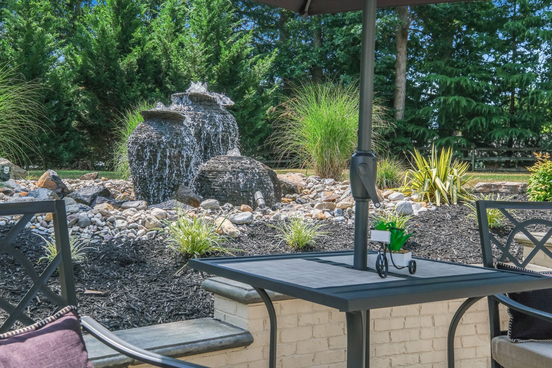 Patio table and chairs facing a stone fountain in a leafy garden area