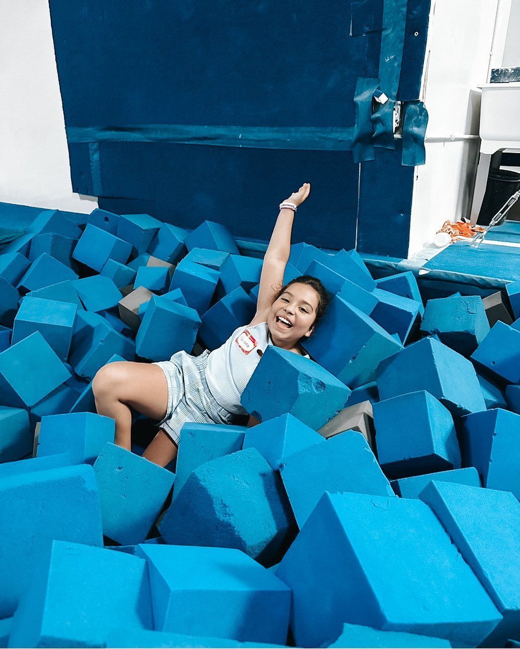 Person smiling in a blue foam pit, arm raised, surrounded by blue foam cubes