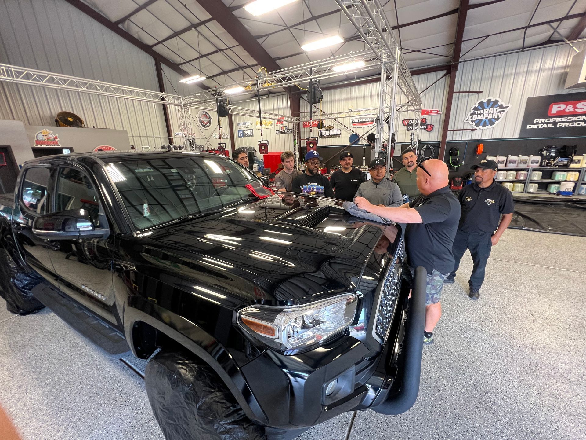 Group of men gathered around a black truck inside a shop. Some are looking at the hood being worked on.