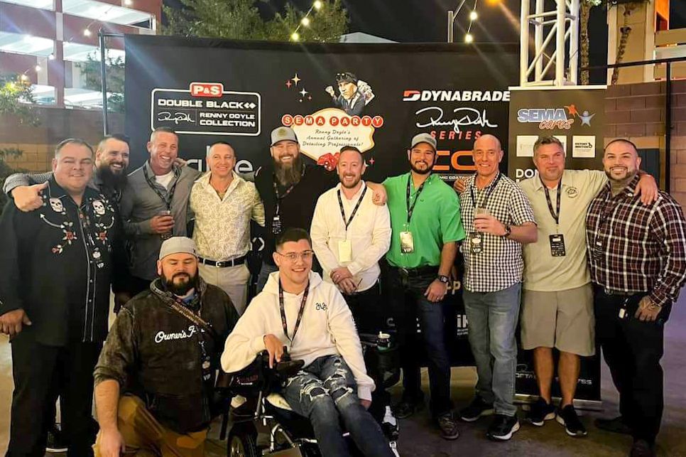 Group of men posing for a photo, some with arms around each other, at an event with a banner backdrop.