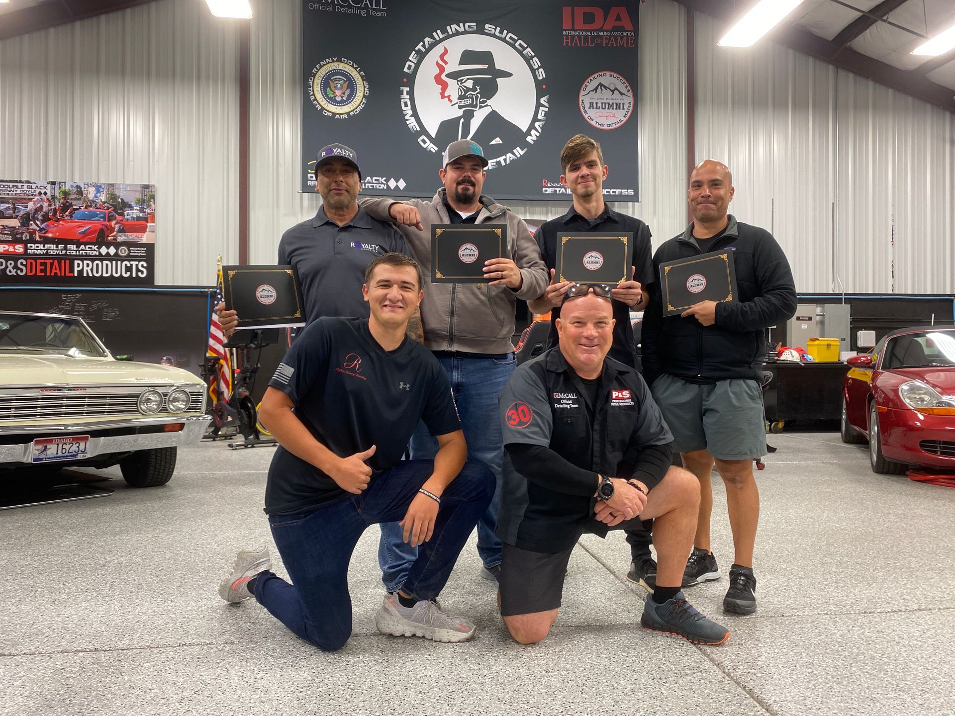Group of men in a garage, holding certificates, smiling at the camera.