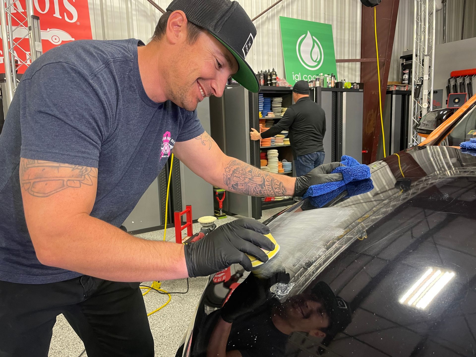 A man wearing a black cap and gloves detailing a car's windshield, smiling. Another man works in the background.