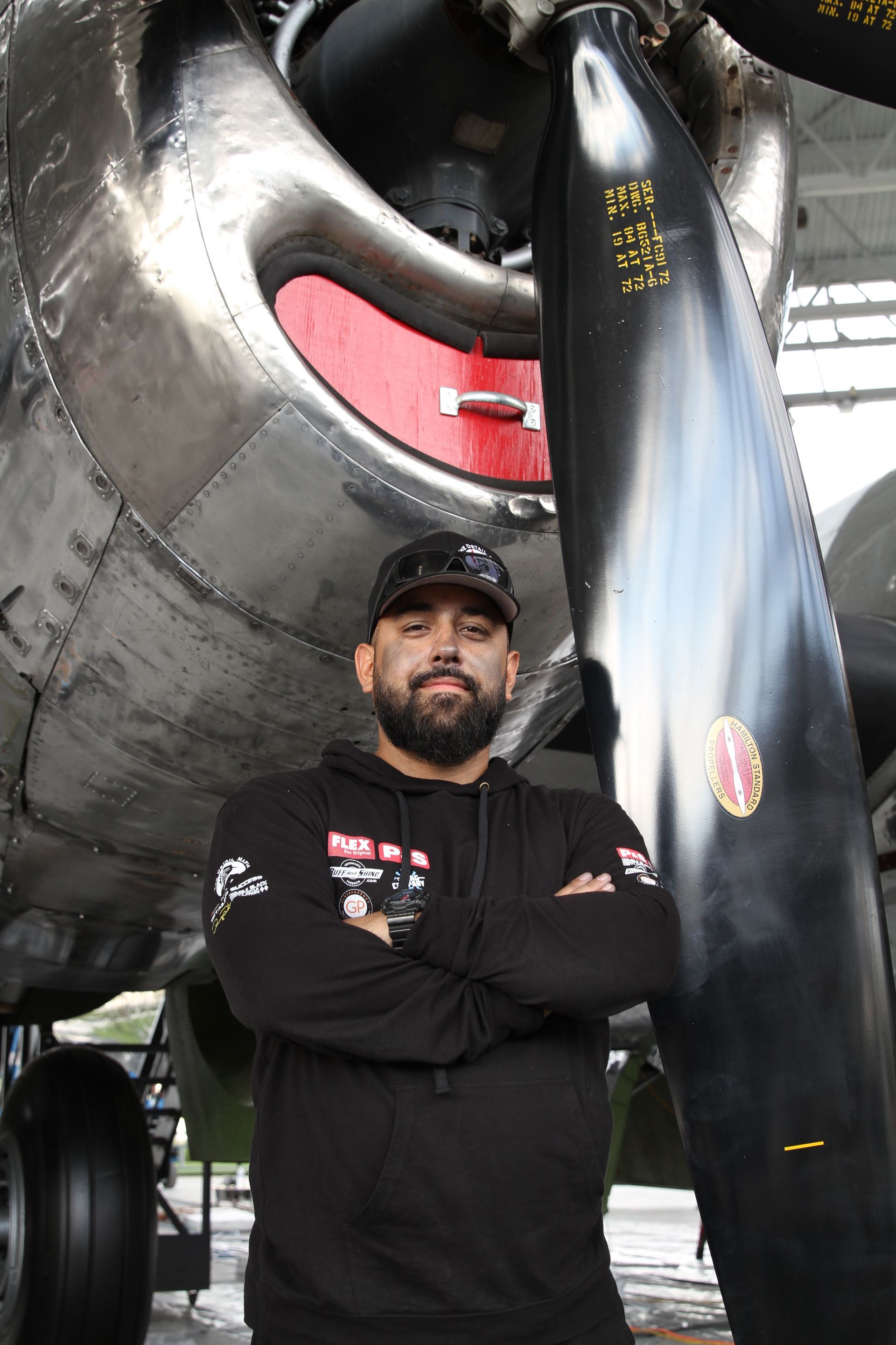 Man with arms crossed in front of a vintage airplane propeller; black hoodie, outdoor hangar.