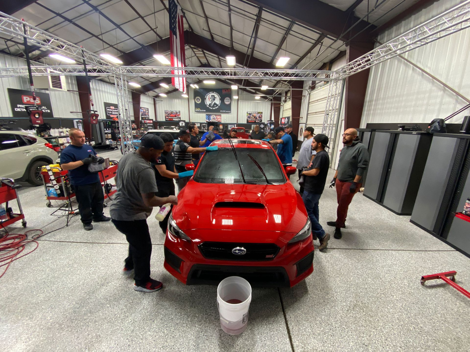 People cleaning a bright red car in a garage, with an American flag hanging.