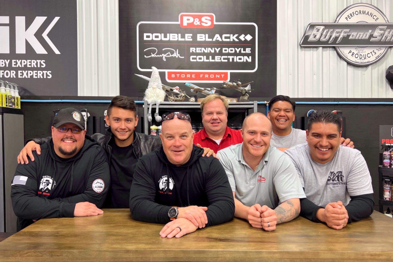 Group of men posing in front of car detailing supplies. They smile, some wearing branded apparel.