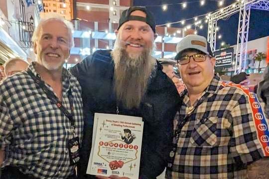 Three men pose outdoors, holding a certificate. Man in the center has a long beard and a baseball cap.