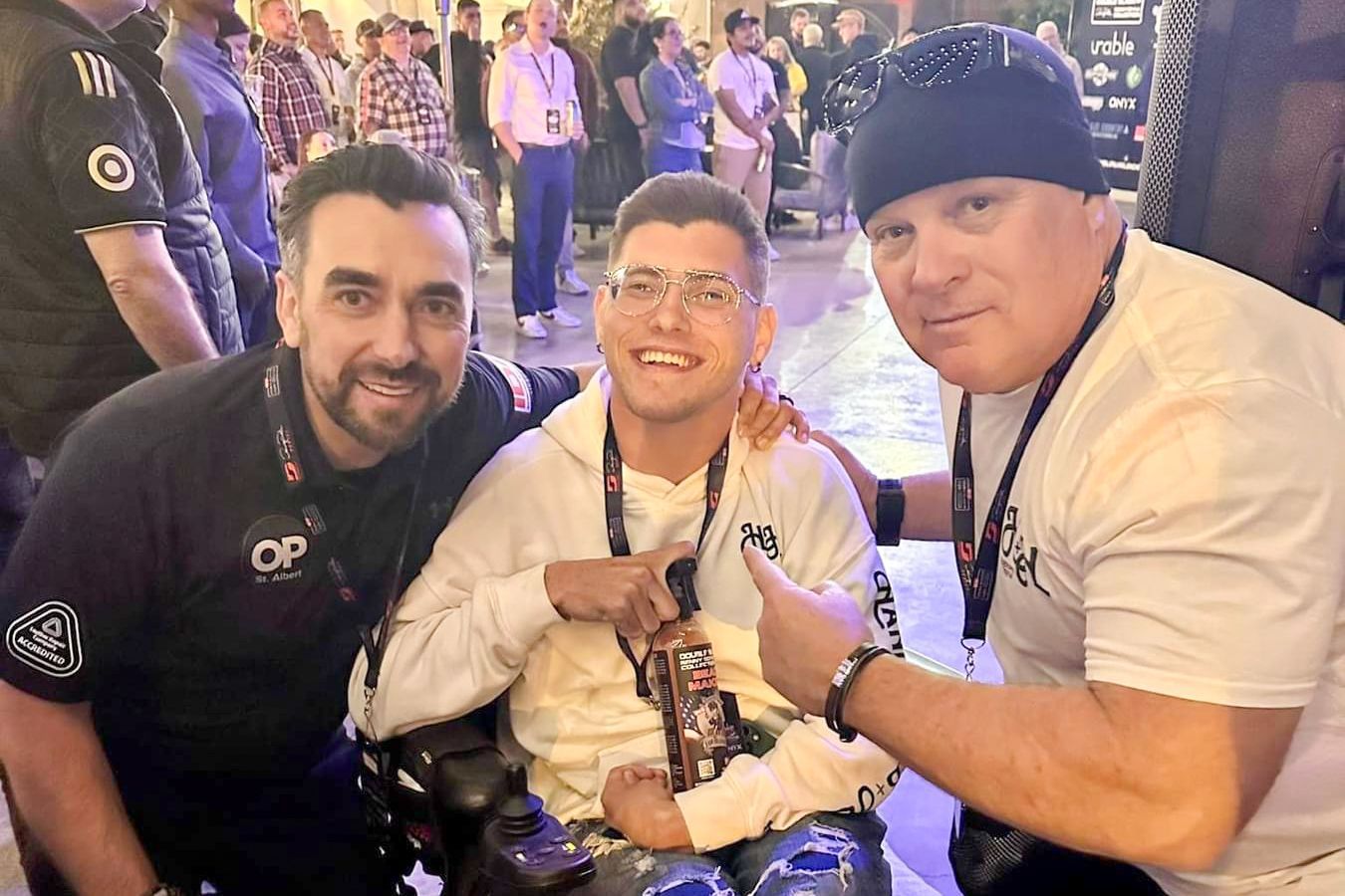 Three men posing for a photo. One in a wheelchair, smiles, gives thumbs up. Others wear black shirts, smile. Outdoors, crowded background.