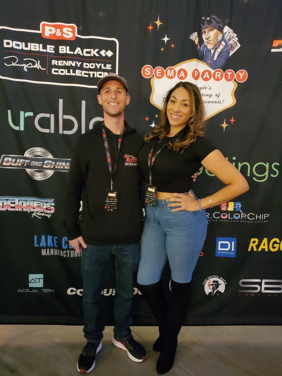 A man and woman pose in front of a banner. The woman wears a black crop top, jeans, and boots. The man wears a hoodie and jeans.