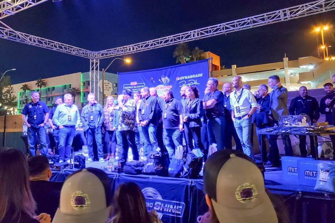 People on stage under lights at an outdoor event, some wearing hats. Nighttime setting.