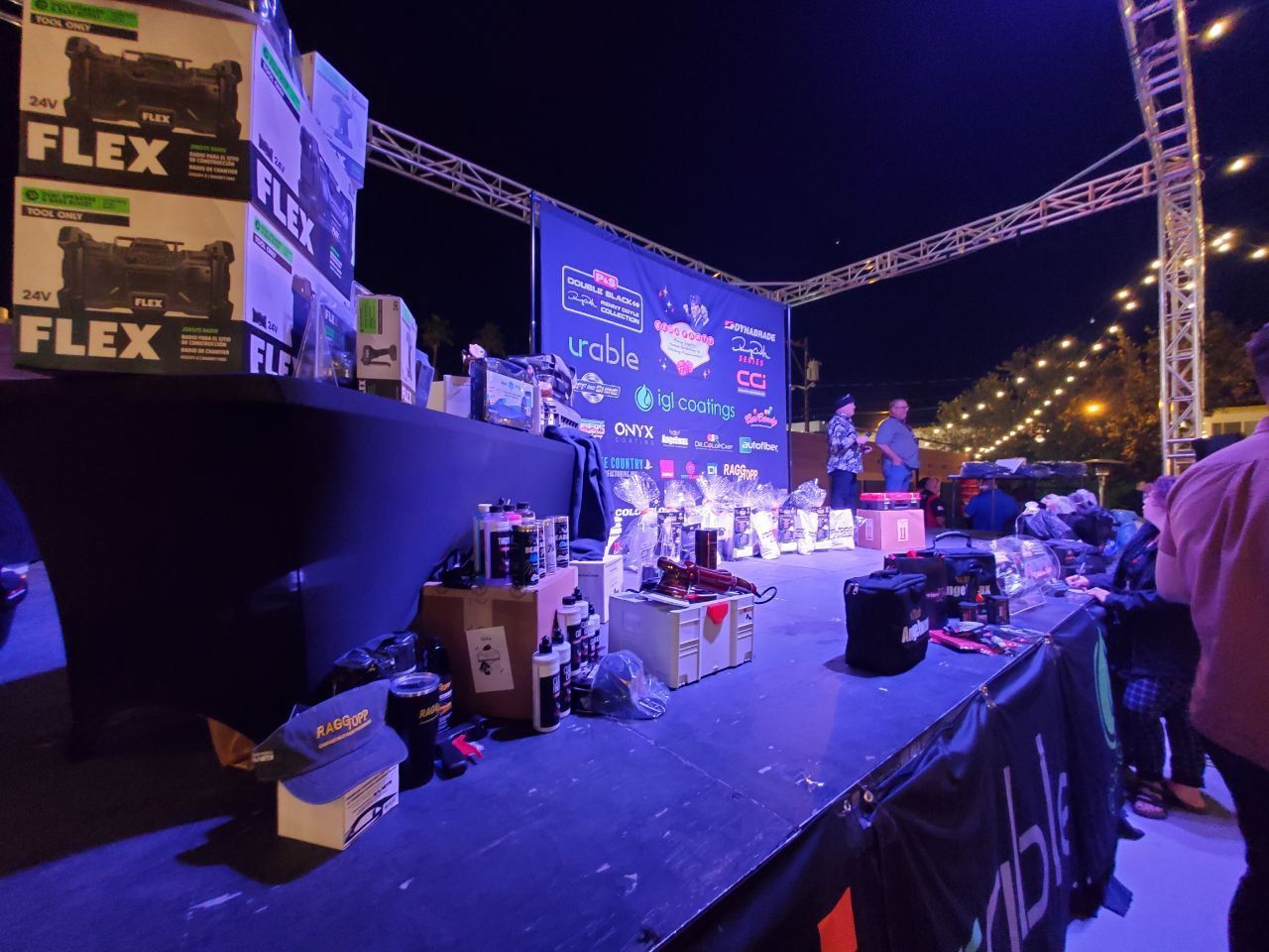 A stage with tools, boxes, and two people; night setting with a sponsor banner, lights, and attendees.