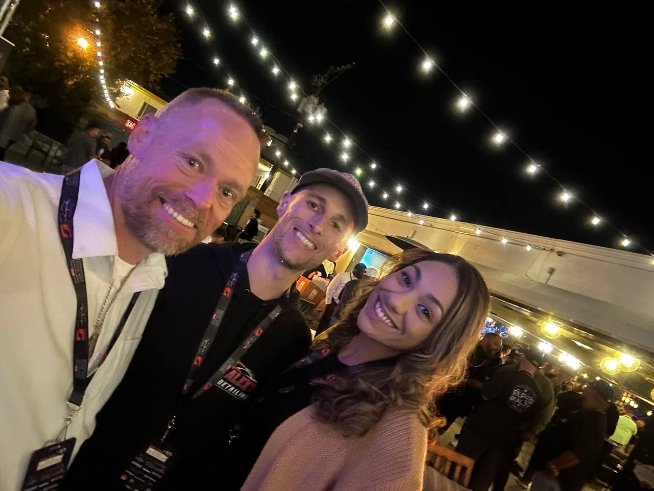 Three smiling people taking a selfie at night under string lights; outdoor event.