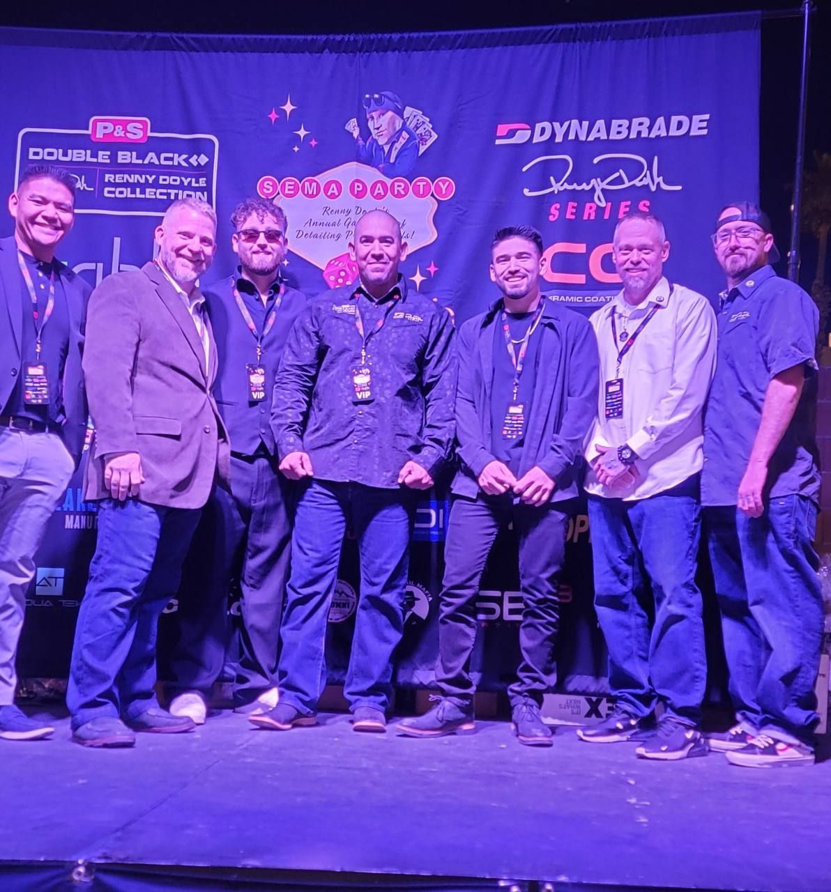 Group of eight men posing on a stage. They are in front of a backdrop that reads 