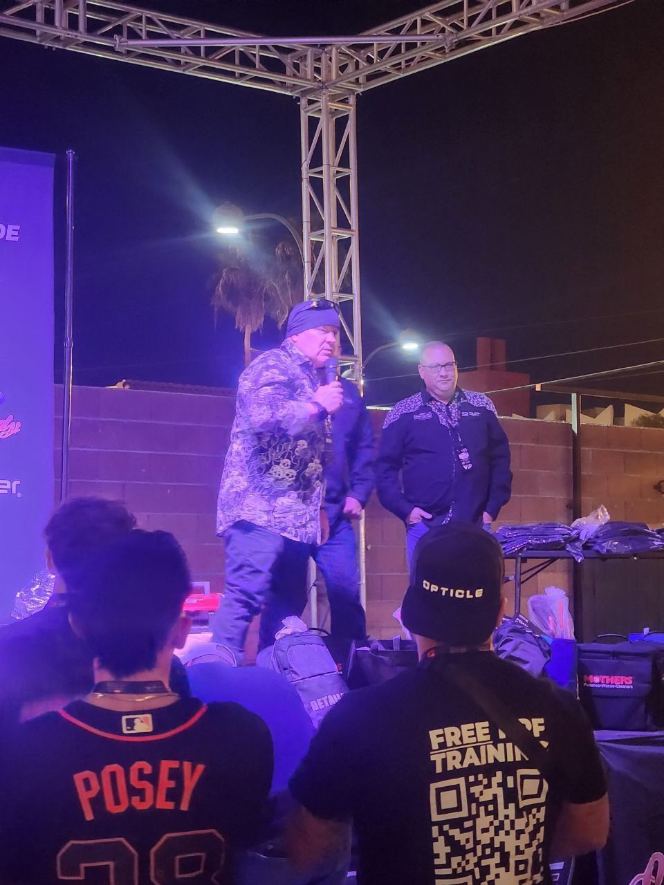 Man in camo jacket speaks into a microphone on a stage, another man stands beside him. Night setting.