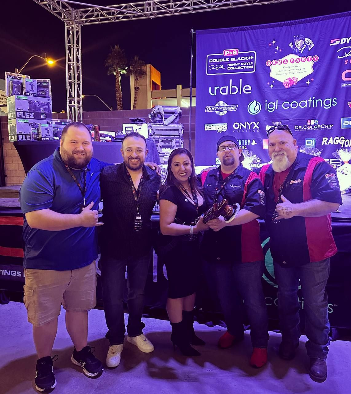 Five people smiling, holding an award on a stage, at a car show outdoors at night.