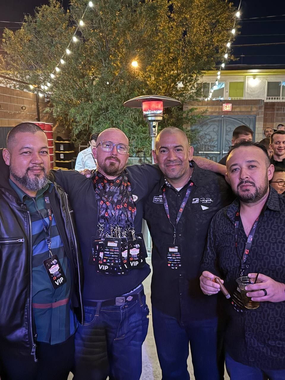 Four men pose outdoors, arms around each other, smiling. Nighttime, string lights.