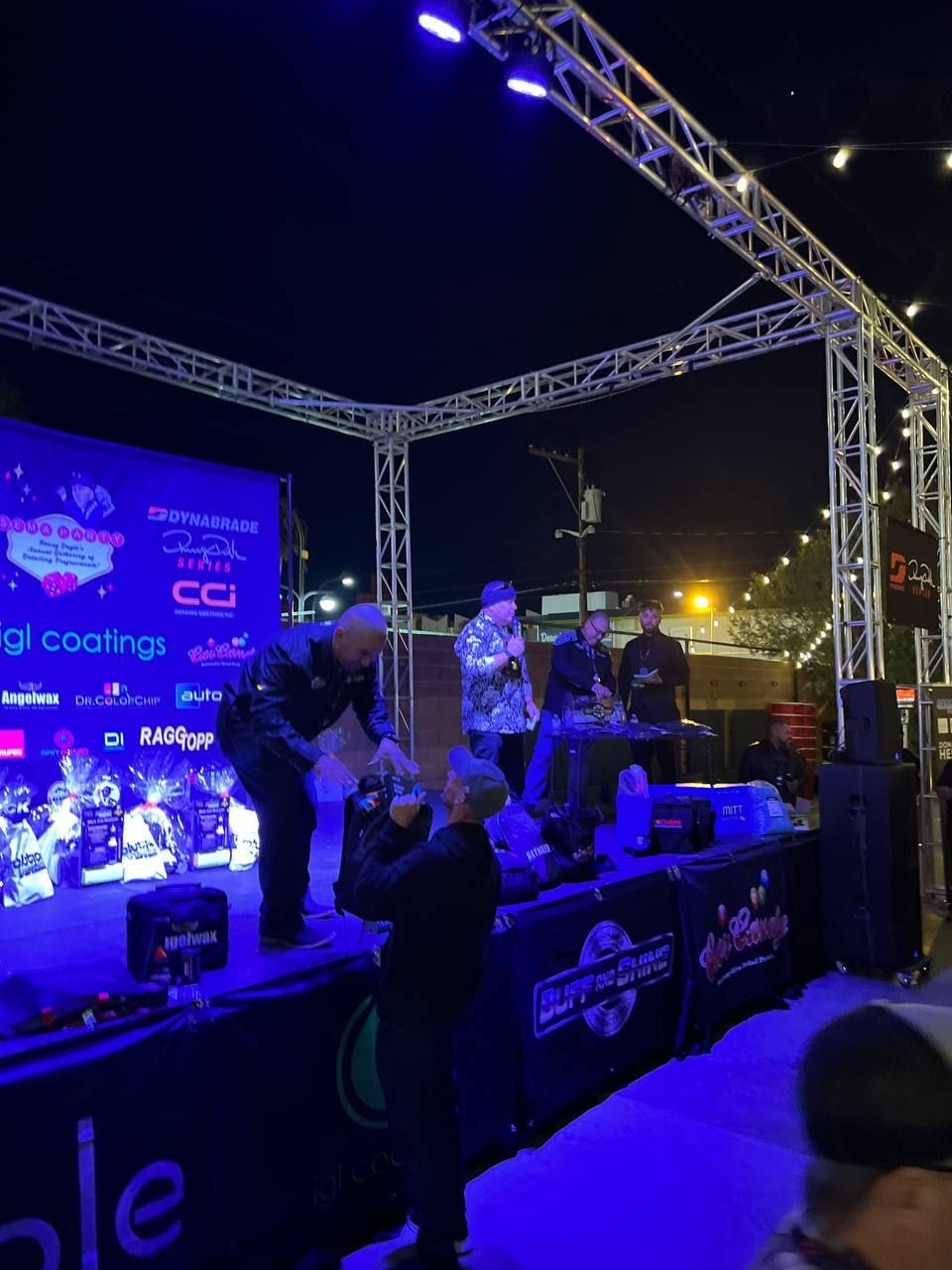 People on a stage under trussing; a contest outdoors at night.