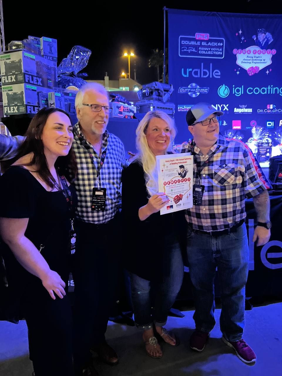 Four people posing, holding a certificate outdoors at night. People smiling, under blue and white lights.
