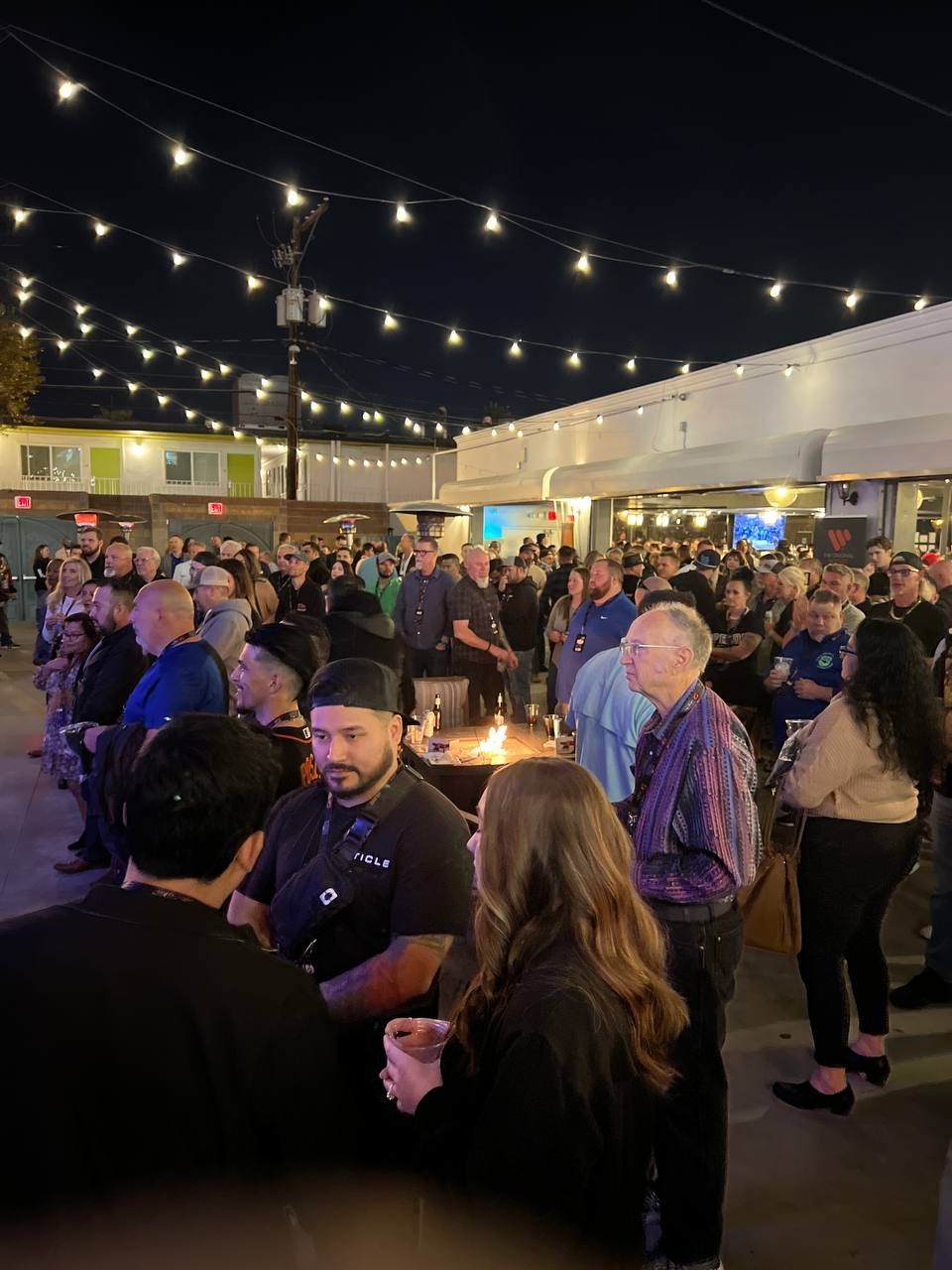 Nighttime outdoor gathering with many people under string lights.