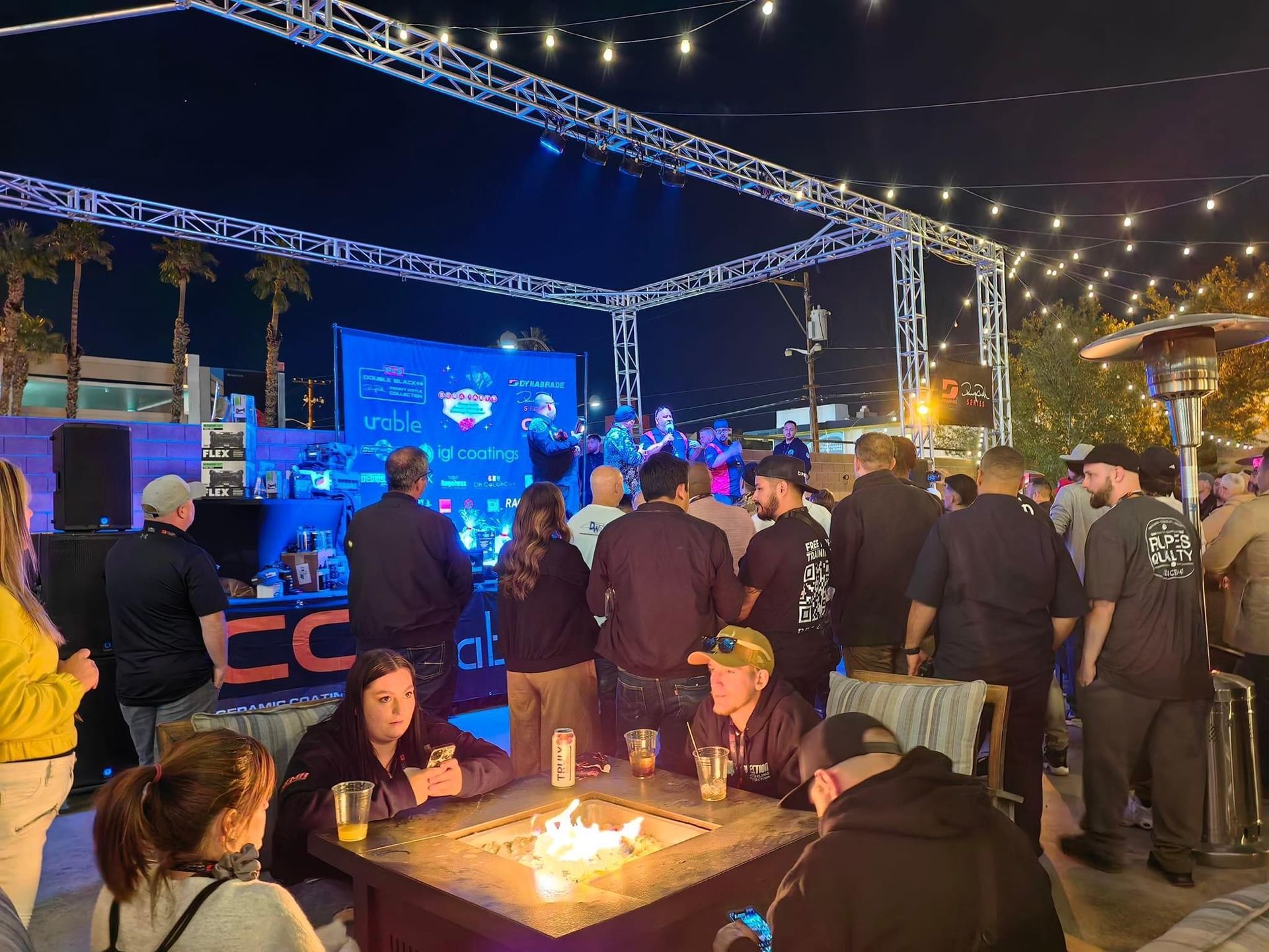Crowd at outdoor event with band on stage, nighttime scene, fire pit, bar area.