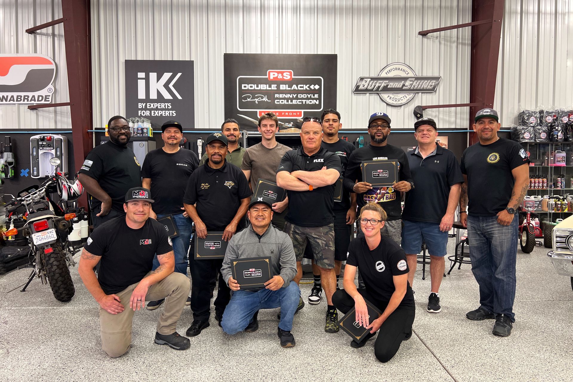 A group of people pose together in an automotive shop. Some hold certificates; smiles and pride are evident.