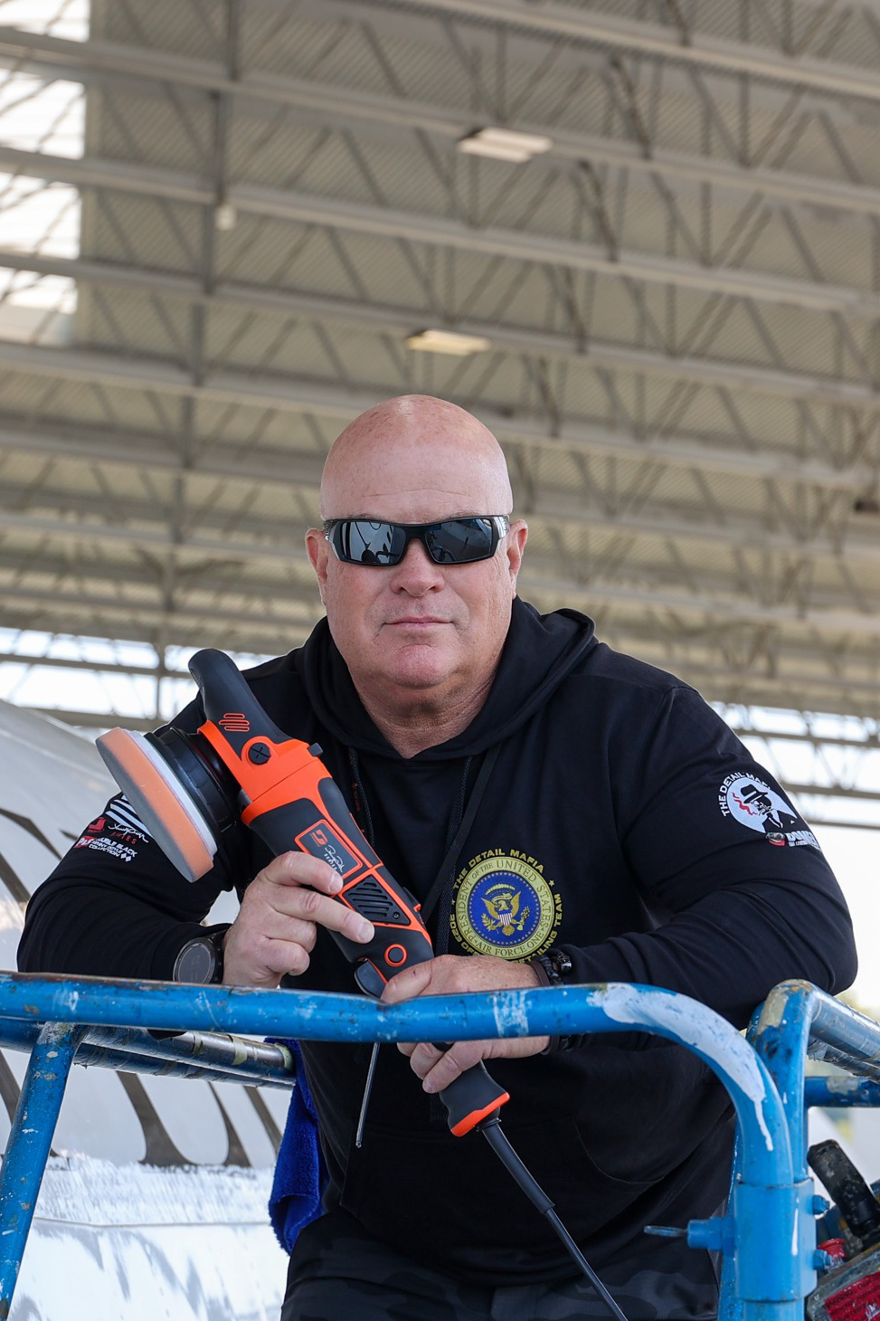 Man in sunglasses holding a polisher, leans on a lift by an aircraft; inside hangar.