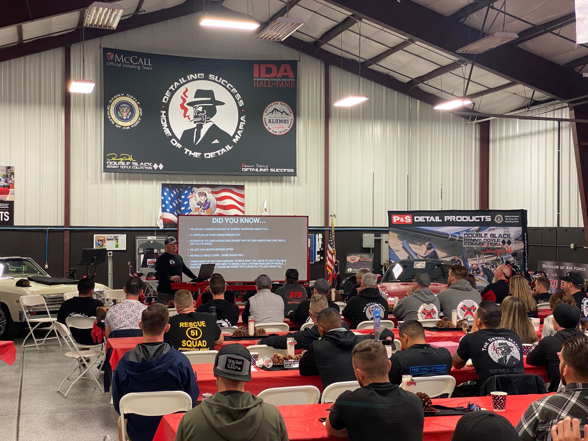Large event: speaker at lectern, audience seated at tables in a hall, presentation screen.