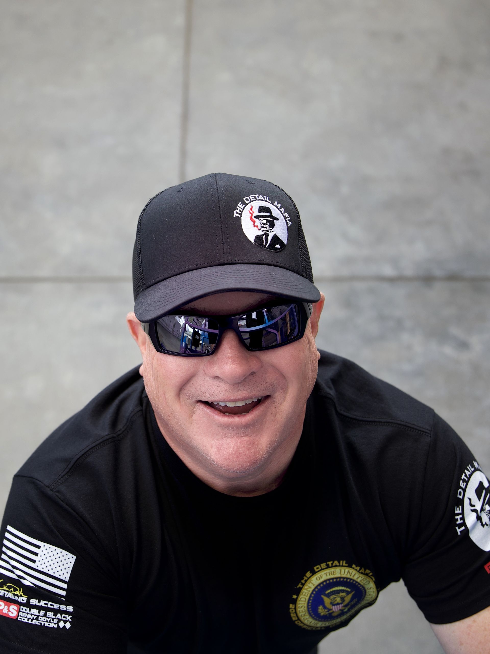 Man in black cap and sunglasses, smiling, wearing a black t-shirt with patches.