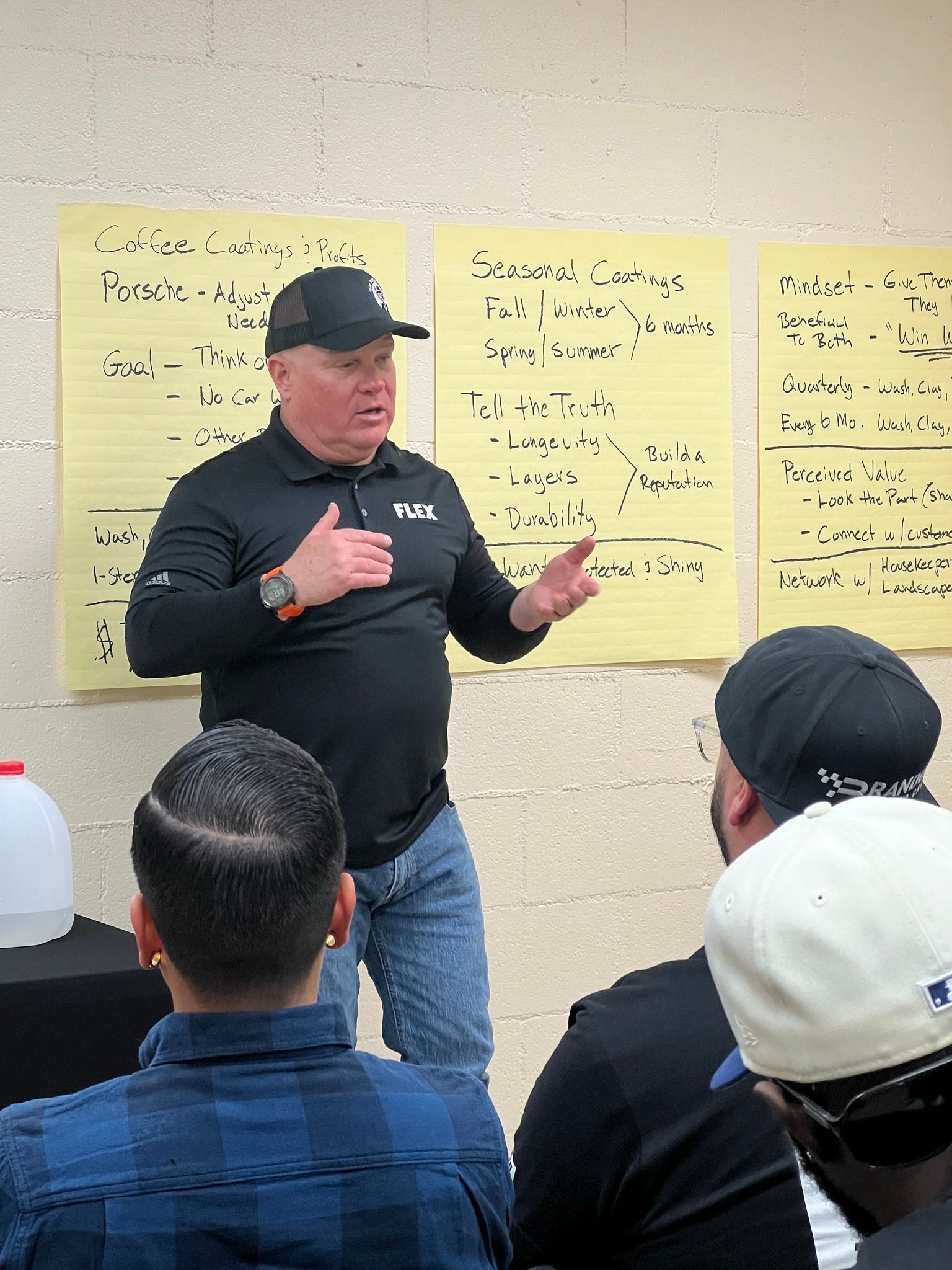 Man in black shirt gestures while speaking to a group. Yellow notes are on a wall behind him.
