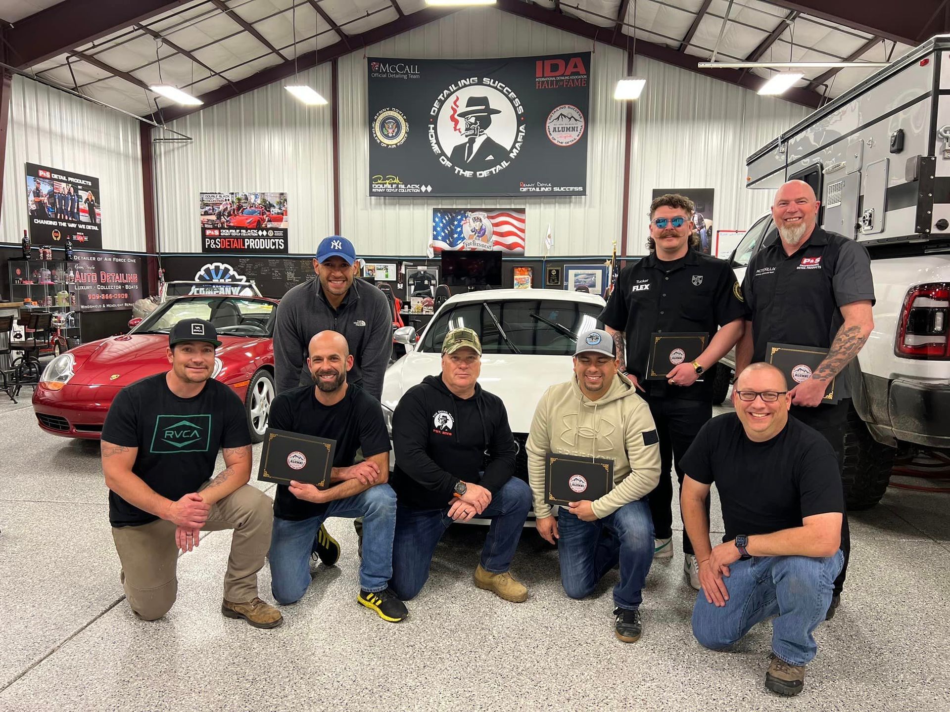A group of men pose with a classic car in a garage; some hold certificates.