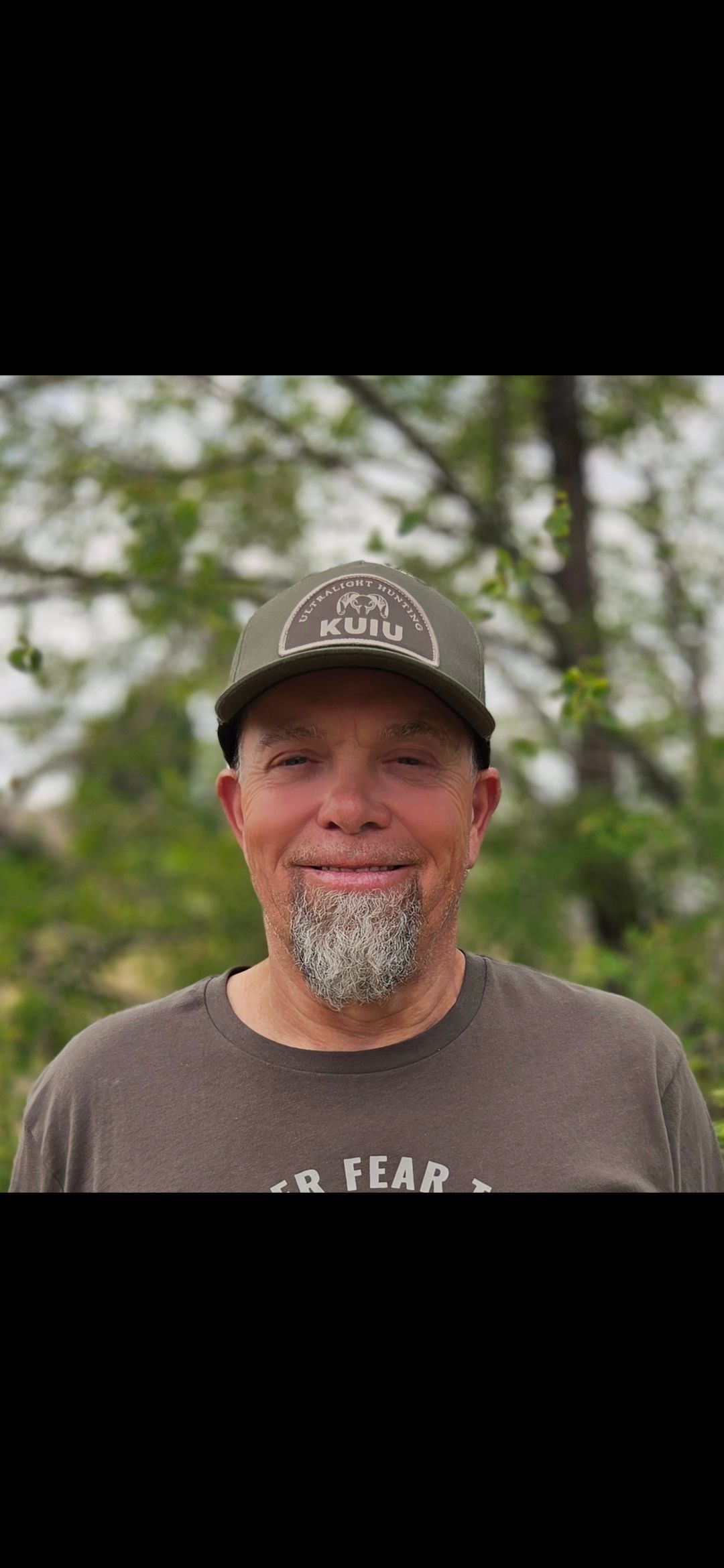 A man with a beard wearing a hat and a t-shirt is smiling.