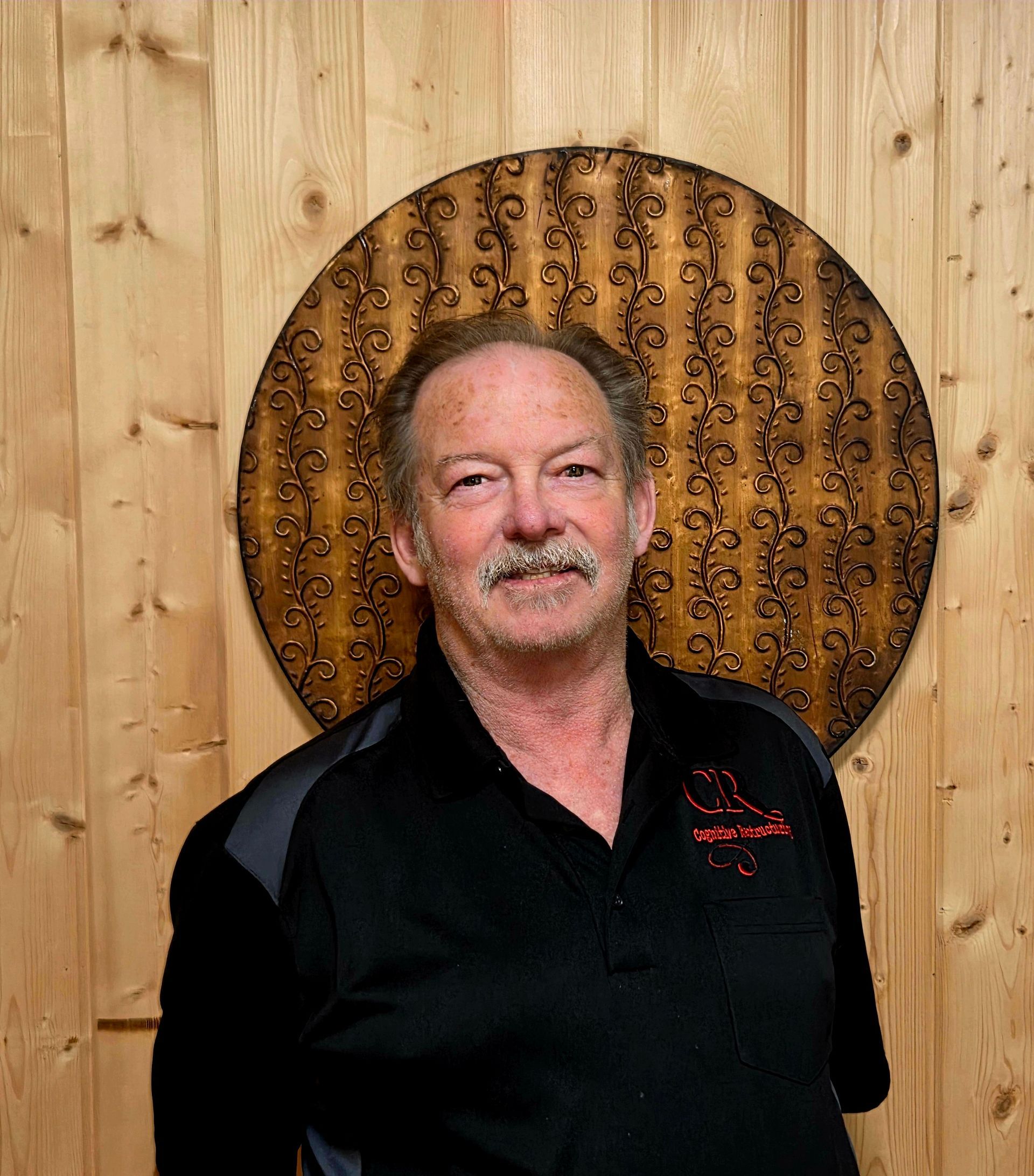 A man with a mustache is standing in front of a wooden wall.