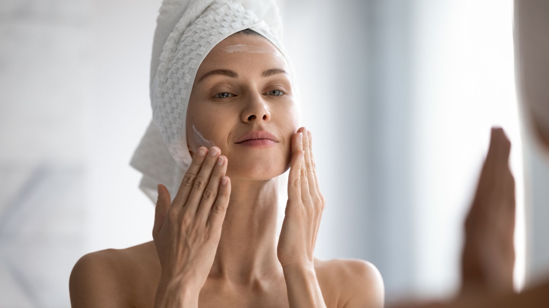 A woman with a towel wrapped around her head is looking at her face in the mirror.