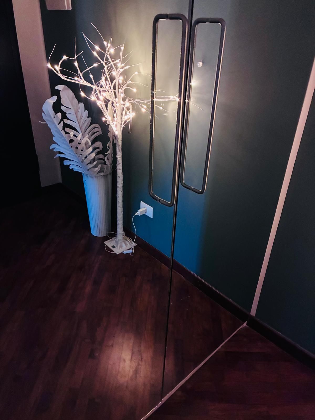 Lampada da terra con due specchi, accanto a un albero decorativo illuminato e un vaso, su un pavimento in legno scuro contro una parete scura.