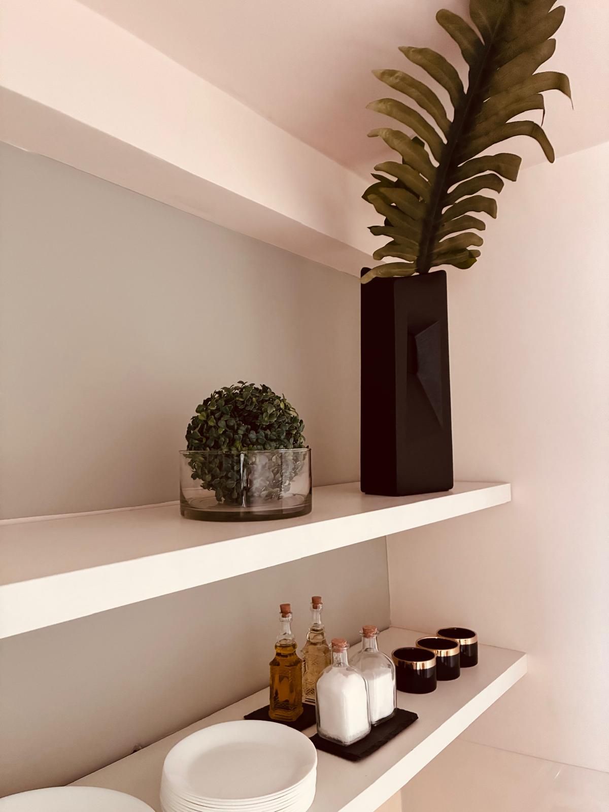 Su mensole bianche sono esposte piante in vaso, bottiglie e candele contro una parete neutra.