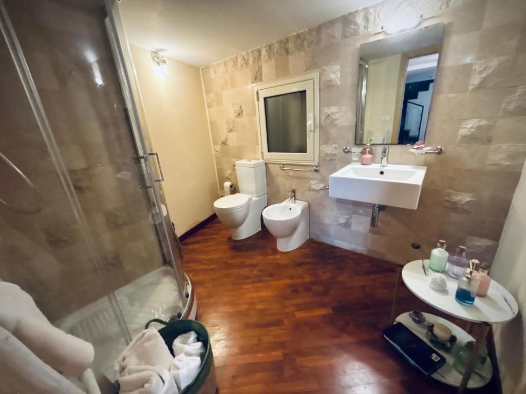 Bagno con WC, bidet, lavandino, specchio e doccia. Pavimento in legno, pareti chiare e un tavolino con oggetti.