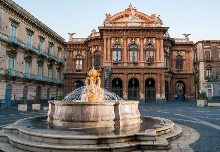 Fontana di fronte a un edificio decorato, di colore rosso-marrone, con ingressi ad arco, forse un teatro, in una piazza lastricata.