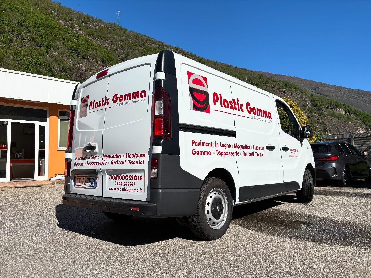 Un furgone commerciale bianco della Plastic Gomma parcheggiato su un piazzale ghiaioso di fronte a un edificio, con le montagne sullo sfondo.