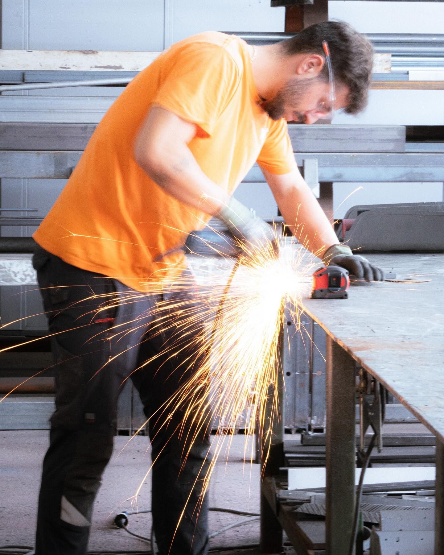 Un operaio con una maglietta arancione usa una smerigliatrice angolare su una lamiera, creando una pioggia di scintille in un'officina.
