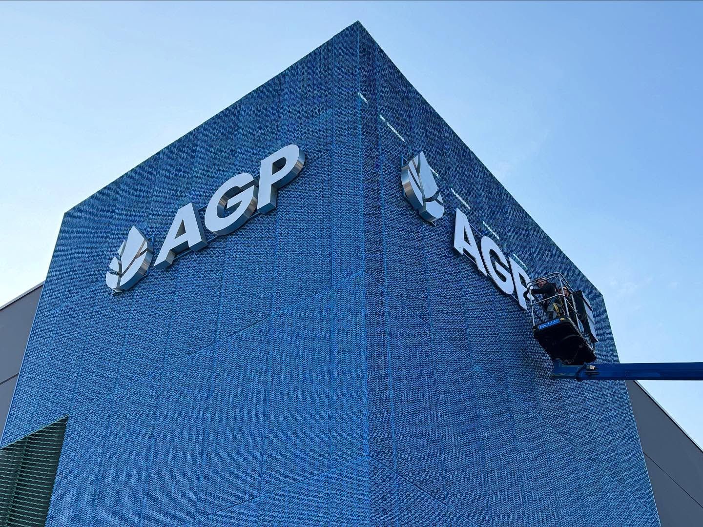 Un operaio, all'interno di un ascensore, installa il logo AGP sulla facciata di un edificio blu dalla superficie ruvida, sullo sfondo di un cielo limpido.