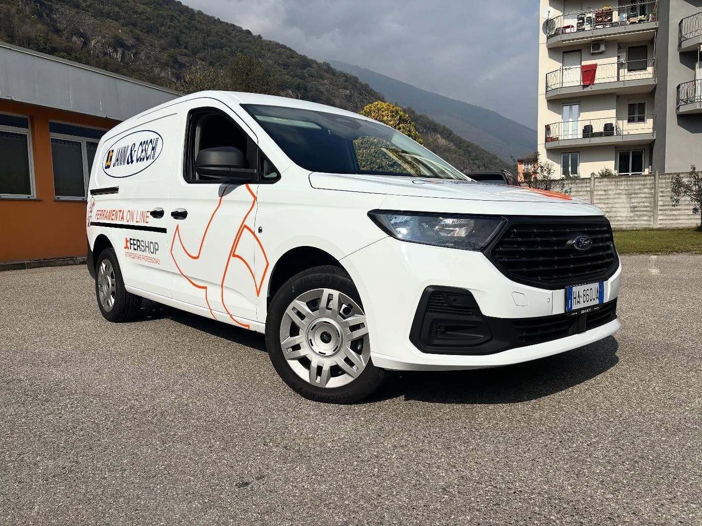 Un furgone per le consegne Ford Transit Connect bianco parcheggiato su un piazzale ghiaioso di fronte a un edificio, con le montagne sullo sfondo.