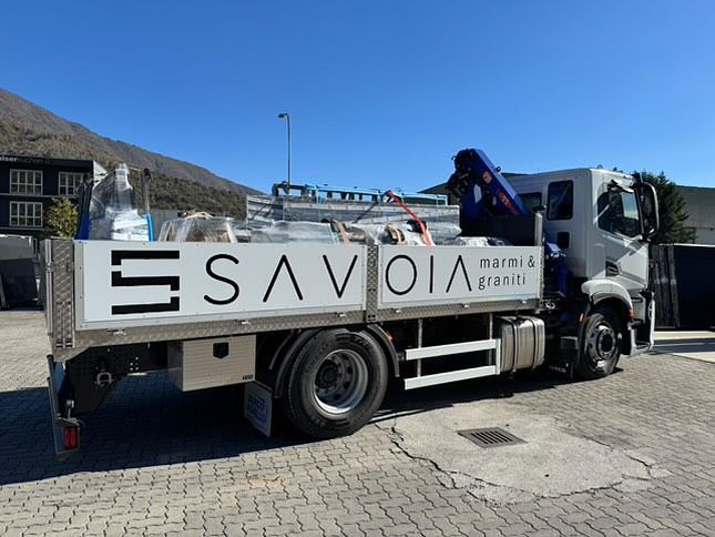 Un camion a pianale ribassato bianco della Savoia Marmi & Graniti con gru montata, parcheggiato su un piazzale asfaltato con le montagne sullo sfondo.