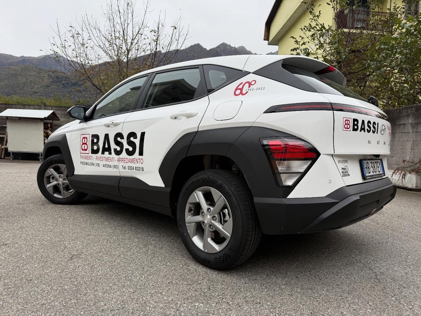 Un SUV Hyundai Kona bianco con il marchio "BASSI", parcheggiato su un piazzale sterrato con le montagne sullo sfondo.
