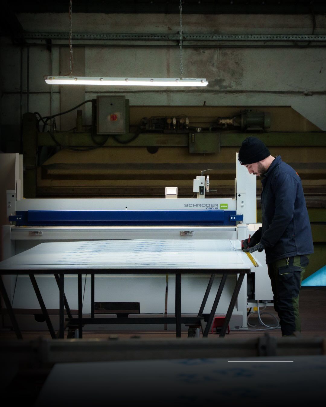 Un operaio con giacca scura e berretto misura un grande foglio bianco su un tavolo di metallo in un'officina con attrezzature industriali.