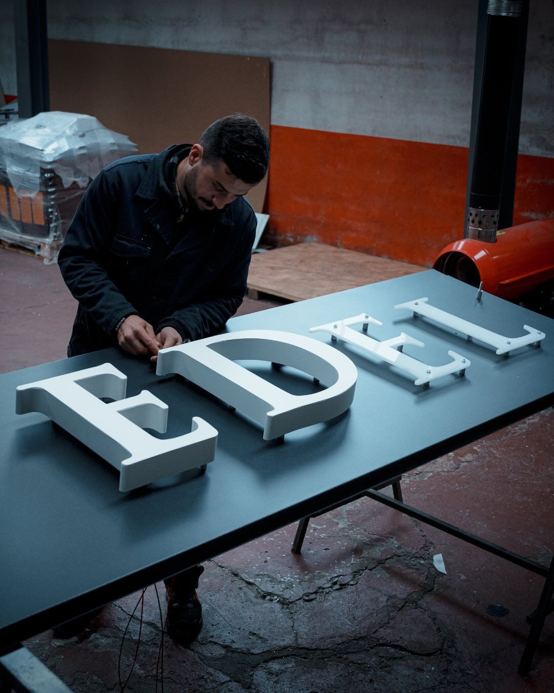 In un'officina, una persona lavora a un banco da lavoro assemblando grandi lettere bianche tridimensionali che compongono la scritta 