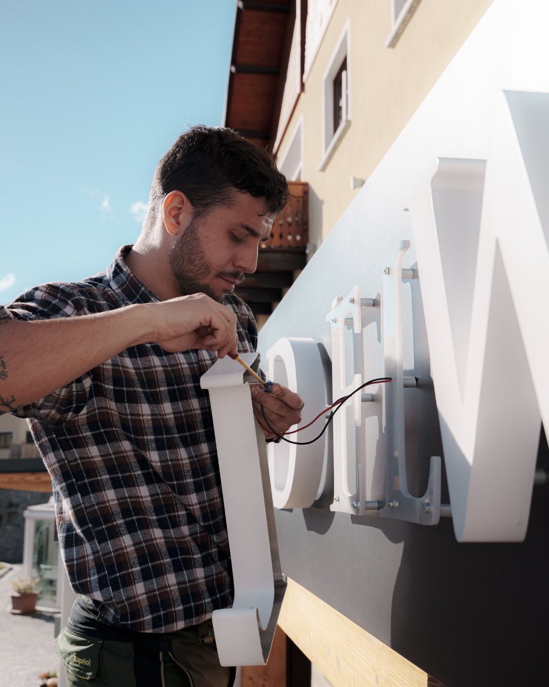 Un tecnico con una camicia a quadri installa dei cavi elettrici in un'insegna tridimensionale bianca su un edificio esterno.