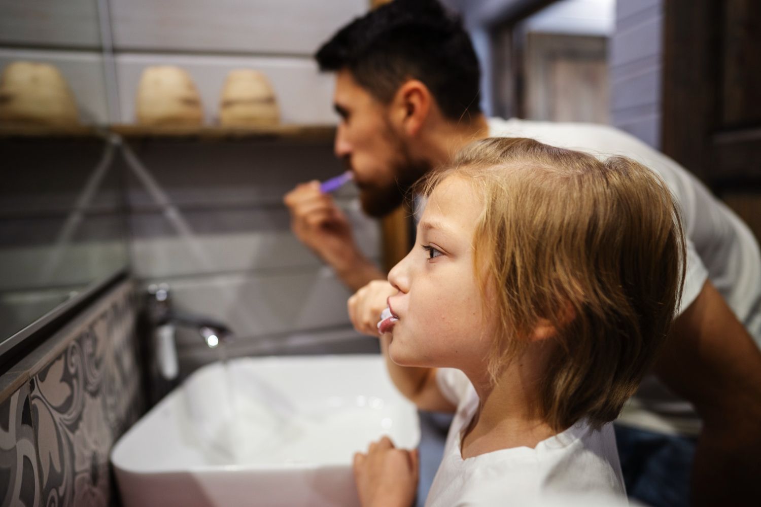 Father and child brushing teeth in a bathroom, facing a mirror.