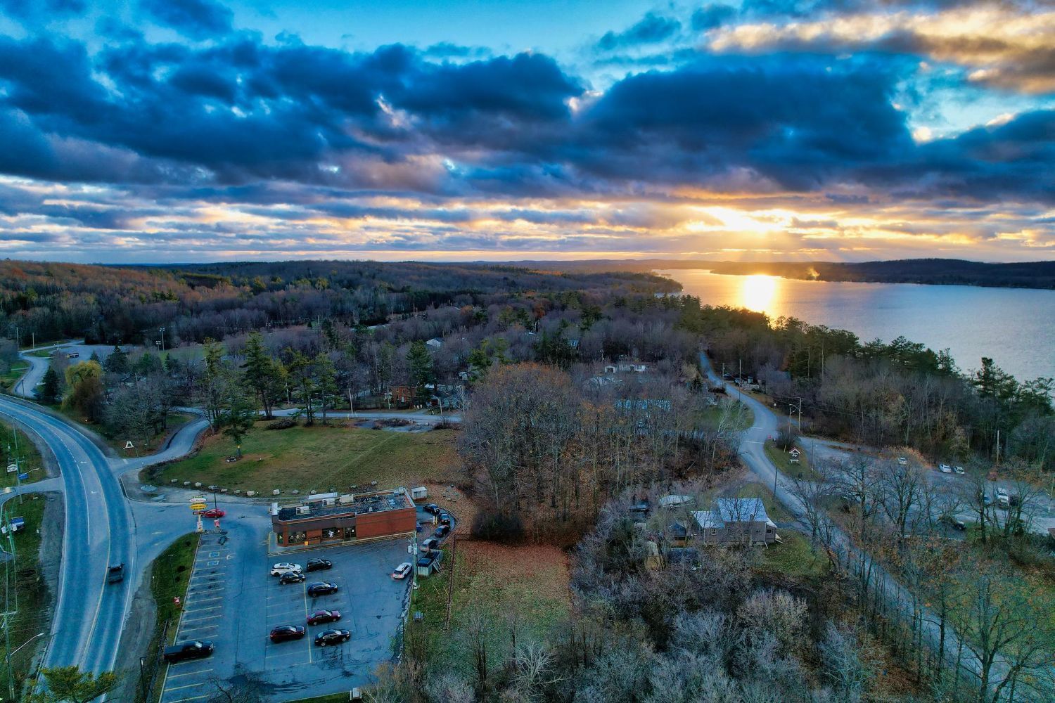 Sunset over a lake and forest. Road, parking lot, and a building are in the foreground.
