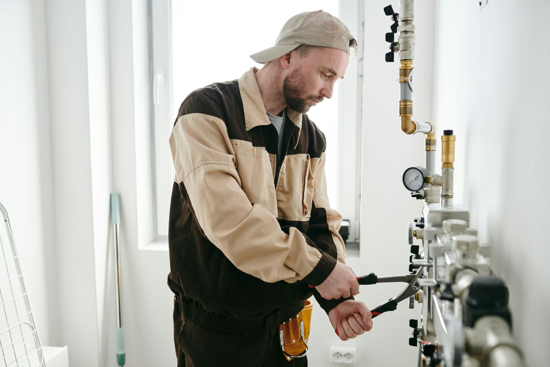 A man in a cap and work clothes, working on pipes with pliers, near a wall.